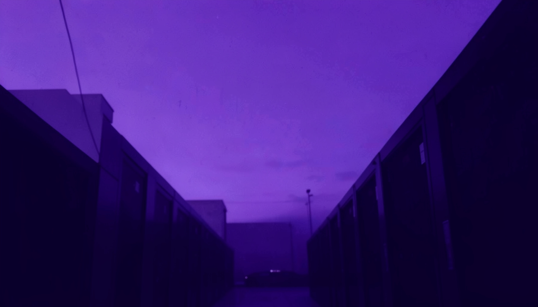 A wide, low-angle duotone photograph of a violet and deep-indigo sky over a faint silhouette of a data-center corridor, rendered as a hard two-tone duotone in lavender and deep purple.